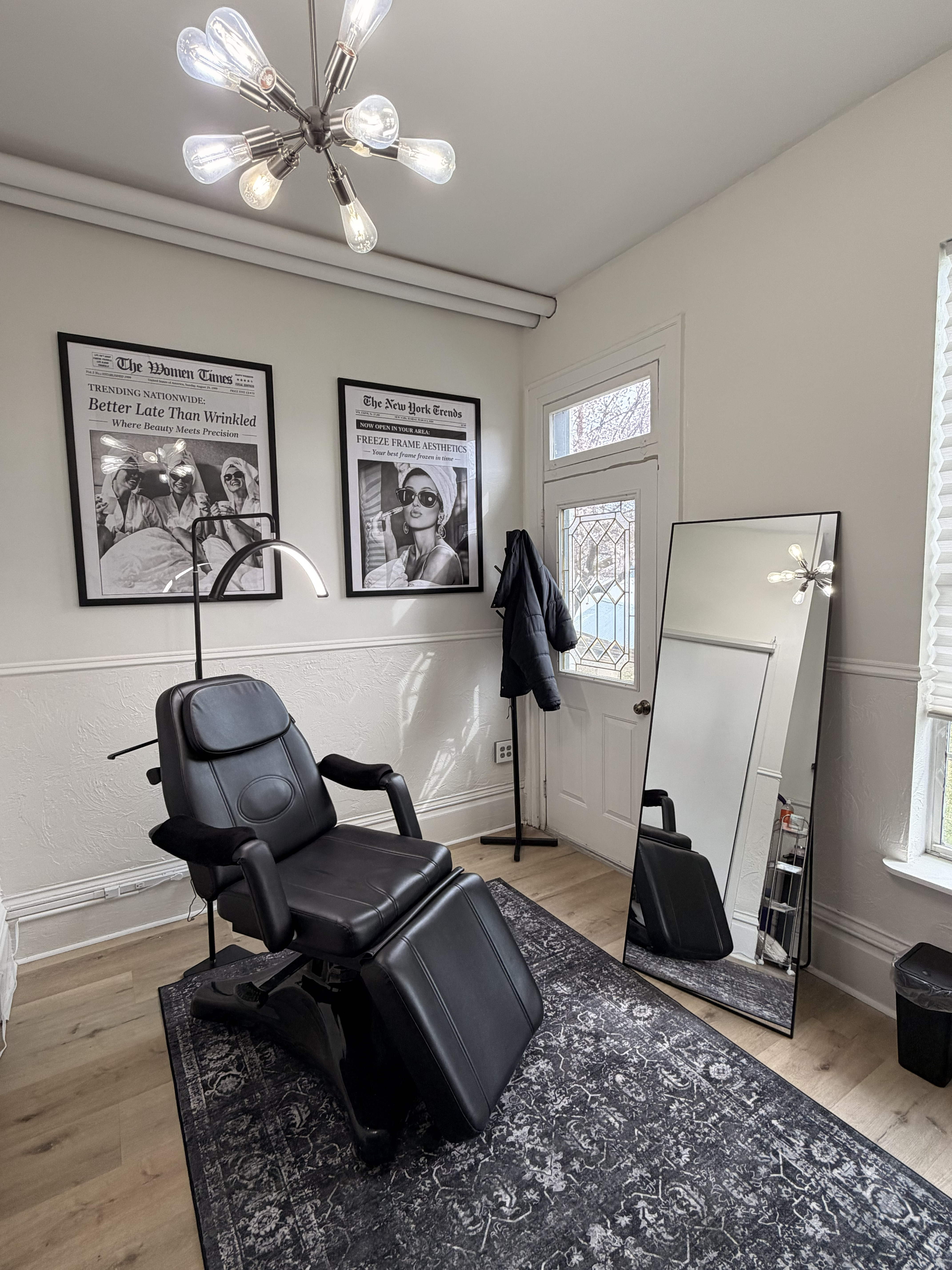 Freeze Frame Aesthetics treatment room — black chair, newspaper art prints, sputnik chandelier, and warm natural light